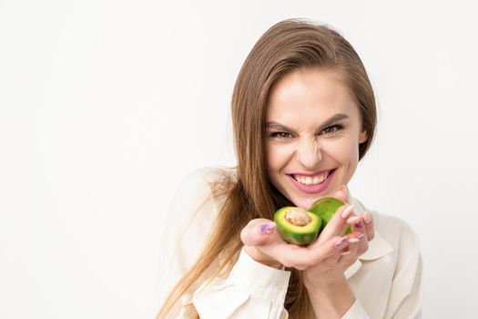 Portrait of a lovely smiling young brunette caucasian woman wearing the white shirt with long hair holding and showing avocado, standing isolated over white background