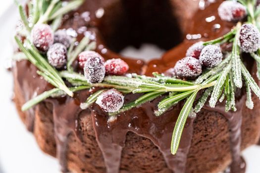 Chocolate bundt cake with chocolate frosting decorated with fresh cranberries and rosemary covered in a white sugar.
