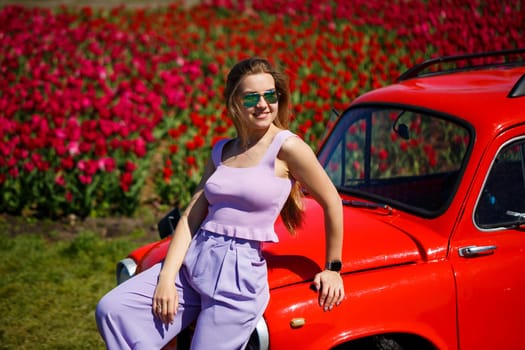 A girl model in casual clothes sits on a red car among tulip fields.