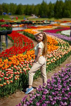 Beautiful woman in hat laughing to camera in spring flower garden. Girl with camera smiling in park with many flowers. Woman enjoying time in blooming flowers field. Pretty lady walking in meadow.