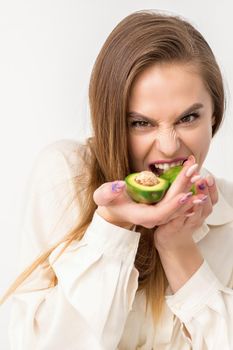 Portrait of a lovely smiling young brunette caucasian woman wearing the white shirt with long hair holding and showing avocado, standing isolated over white background