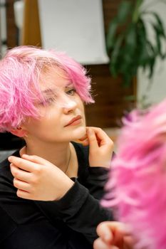 The beautiful young caucasian woman with a new short pink hairstyle looking at her reflection in the mirror checking hairstyle in a hairdresser salon