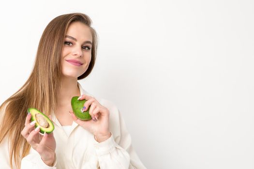 Portrait of a lovely smiling young brunette caucasian woman wearing the white shirt with long hair holding and showing avocado, standing isolated over white background