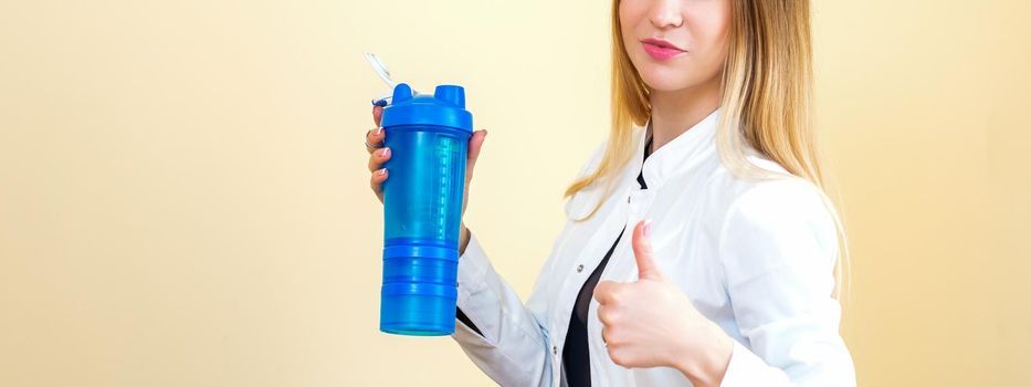 The young Caucasian female doctor wearing a white coat holds a blue plastic bottle with water with thumb up looking at the camera