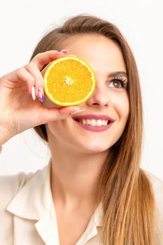 Portrait of a cheerful caucasian young woman covering eye with an orange slice wearing a white shirt over a white background