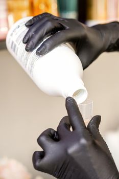 A hairdresser in black gloves is preparing hair dye with a bottle in a hair salon, close up