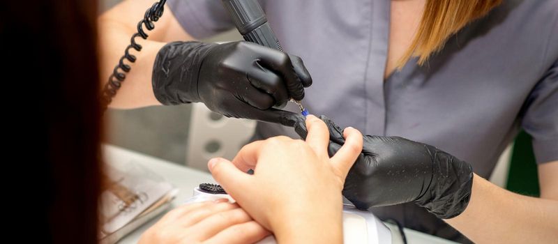 Manicure master in rubber gloves applies an electric nail file to remove the nail polish in a nail salon