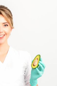 Portrait of smiling young female nutritionist doctor with organic avocado fruits posing at camera on white background, copy space. Benefits of proper nutrition