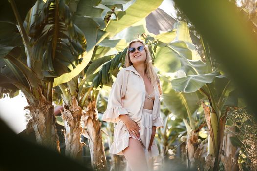 A beautiful young woman with long blond hair of European appearance stands near the banana trees. Girl in tropical forest on a sunny summer day