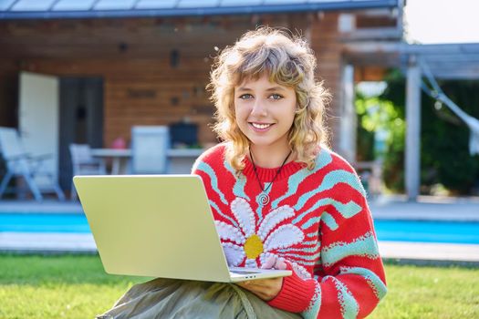 Portrait of teenage girl with laptop outdoor, smiling cute female teenager posing looking at camera, sitting on backyard lawn. Beautiful girl 16, 17 years old with curly hair, high school student