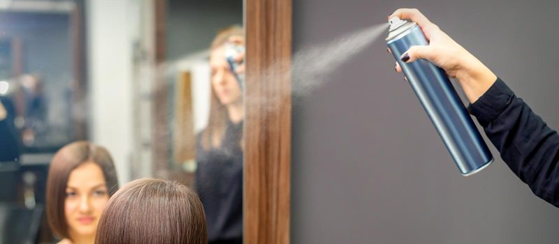 A hairdresser is using hair spray to fix the short hairstyle of the young brunette woman sitting in the hair salon