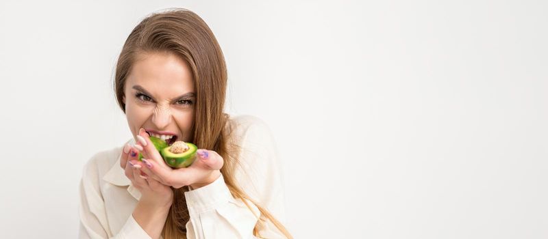 Portrait of a lovely smiling young brunette caucasian woman wearing the white shirt with long hair holding and showing avocado, standing isolated over white background