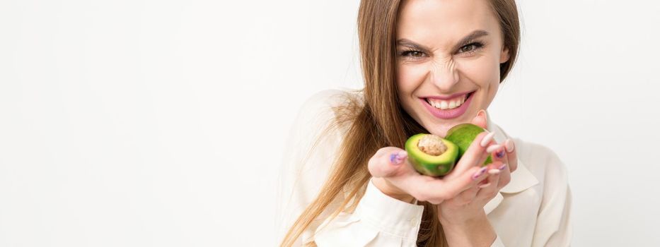 Portrait of a lovely smiling young brunette caucasian woman wearing the white shirt with long hair holding and showing avocado, standing isolated over white background