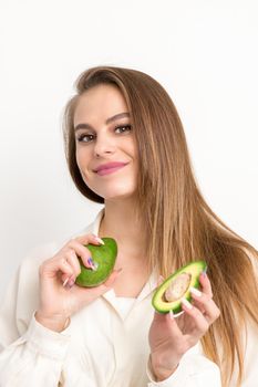 Portrait of a lovely smiling young brunette caucasian woman wearing the white shirt with long hair holding and showing avocado, standing isolated over white background