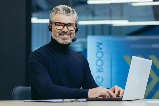 Portrait of mature gray haired businessman, man in glasses smiling and looking at camera, senior boss with headset for video call using laptop, executive working inside modern office building.