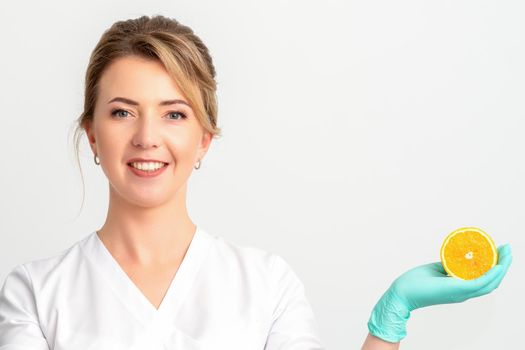 Smiling female nutritionist holding a sliced orange, looking at camera over white background, healthy diet concept
