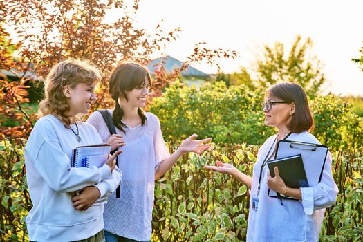 Female teacher talking to teenage female students outdoor. Two girls 17, 18 years old with middle age mentor. Teaching, education, youth, high school, college, university concept