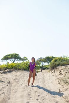 A slender tanned young woman in a swimsuit and a pareo, standing on the golden sand in the desert. Model woman resting and sunbathing on vacation. Travel concept, swimwear
