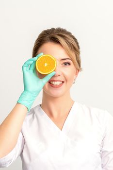 Portrait of young caucasian smiling female beautician covering eye with an orange slice wearing gloves over a white background