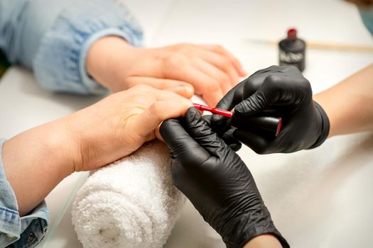 Manicure varnish painting. Close-up of a manicure master wearing rubber black gloves applying red varnish on a female fingernail in the beauty salon