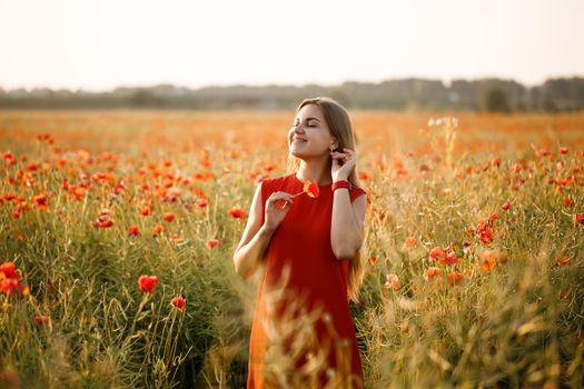 A woman of European appearance with long blonde hair and a red summer dress, she stands in a blooming poppy field
