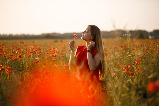 A woman of European appearance with long blonde hair and a red summer dress, she stands in a blooming poppy field