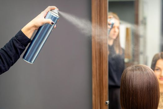 A hairdresser is using hair spray to fix the short hairstyle of the young brunette woman sitting in the hair salon