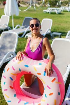 A slender young woman holds an inflatable circle in her hands, standing on the green grass. Attractive model with perfect body. Vacation concept, girl goes swimming in the sea