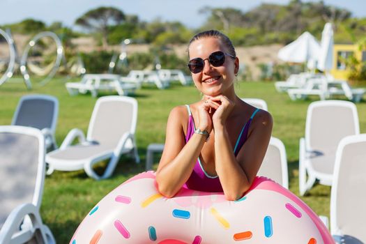 A young woman in a stylish swimsuit and sunglasses holds an inflatable donut ring in her hand. Beautiful happy girl posing and having fun in the sun. Vacation. travel