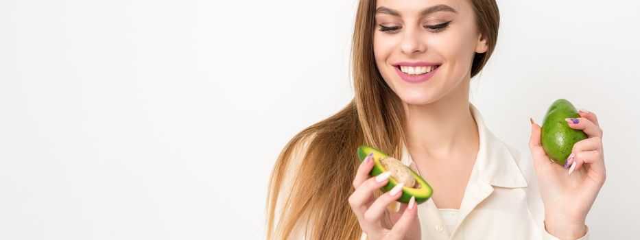 Portrait of a lovely smiling young brunette caucasian woman wearing the white shirt with long hair holding and showing avocado, standing isolated over white background