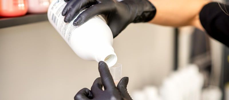 A hairdresser in black gloves is preparing hair dye with a bottle in a hair salon, close up