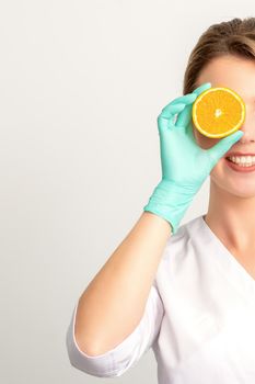 Portrait of young caucasian smiling female beautician covering eye with an orange slice wearing gloves over a white background