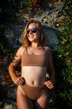 Beautiful girl in sunglasses in a beige swimsuit. Young woman with tanned body soit near stone wall with green plants