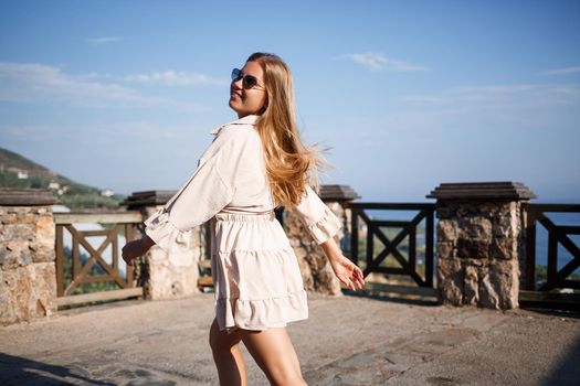 A young woman with long blond hair in a top and a skirt is walking on a summer sunny day. Happy girl with a smile on her face and in sunglasses is walking on a sunny day
