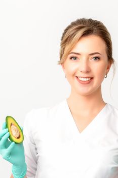 Portrait of smiling young female nutritionist doctor with organic avocado fruits posing at camera on white background, copy space. Benefits of proper nutrition