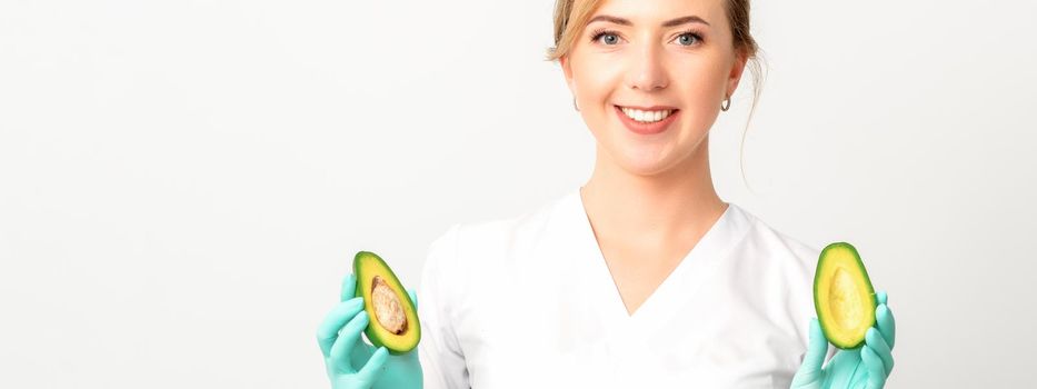 Portrait of smiling young female nutritionist doctor with organic avocado fruits posing at camera on white background, copy space. Benefits of proper nutrition