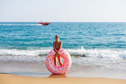 Attractive young woman in a bright swimsuit posing on the beach with an inflatable ring. Beautiful blond woman with long hair relaxing by the ocean. The concept of a sports model, swimwear