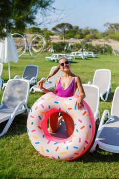 A young woman in a stylish swimsuit and sunglasses holds an inflatable donut ring in her hand. Beautiful happy girl posing and having fun in the sun. Vacation. travel