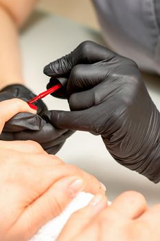 Manicure varnish painting. Close-up of a manicure master wearing rubber black gloves applying red varnish on a female fingernail in the beauty salon