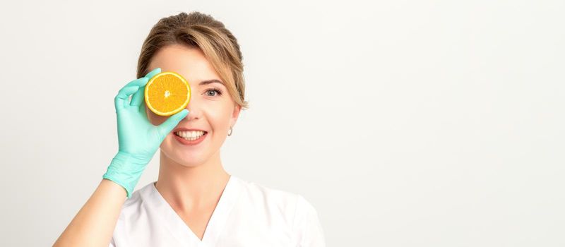 Portrait of young caucasian smiling female beautician covering eye with an orange slice wearing gloves over a white background