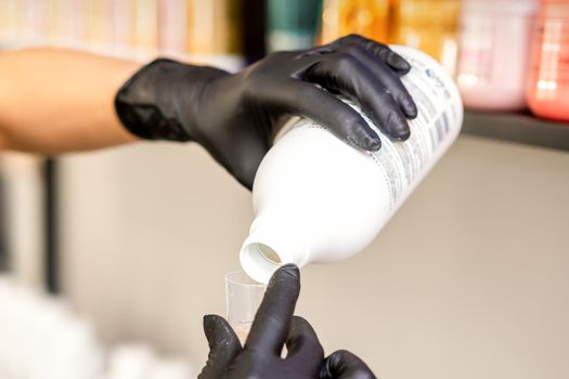 A hairdresser in black gloves is preparing hair dye with a bottle in a hair salon, close up