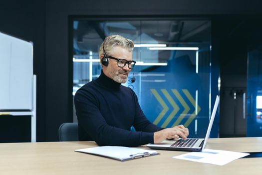 Senior gray haired investor businessman working inside modern office using laptop and headset for video call and remote meeting with colleagues and partners.
