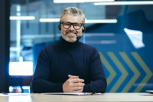Webcam view, senior experienced gray haired businessman looking at camera and smiling, online meeting, video call man with headset working inside modern office building.