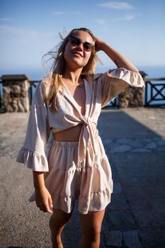 A young woman with long blond hair in a top and a skirt is walking on a summer sunny day. Happy girl with a smile on her face and in sunglasses is walking on a sunny day