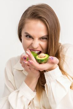 Portrait of a lovely smiling young brunette caucasian woman wearing the white shirt with long hair holding and showing avocado, standing isolated over white background