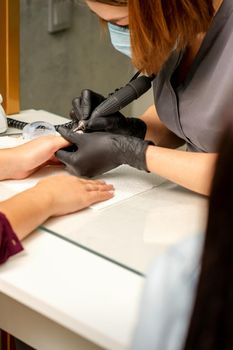 Manicure master wearing protective mask uses electric nail file machine in a nail salon, close up