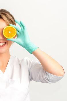 Portrait of young caucasian smiling female beautician covering eye with an orange slice wearing gloves over a white background