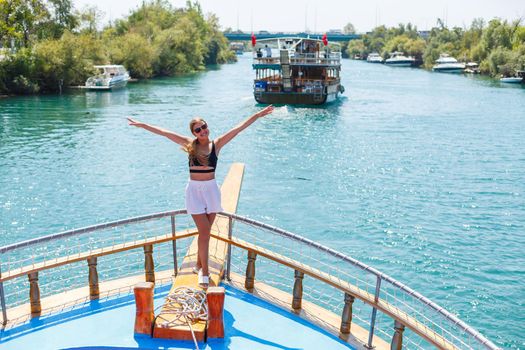 A young slender tanned woman in white shorts and a top, wearing sunglasses. Walk on a hot sunny day on a yacht along the river