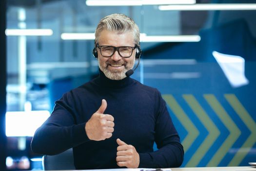 Video call senior gray haired man talking to colleagues remotely using headset smiling and looking at camera giving thumbs up, mature businessman inside modern office.