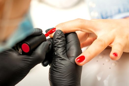 Manicure varnish painting. Close-up of a manicure master wearing rubber black gloves applying red varnish on a female fingernail in the beauty salon